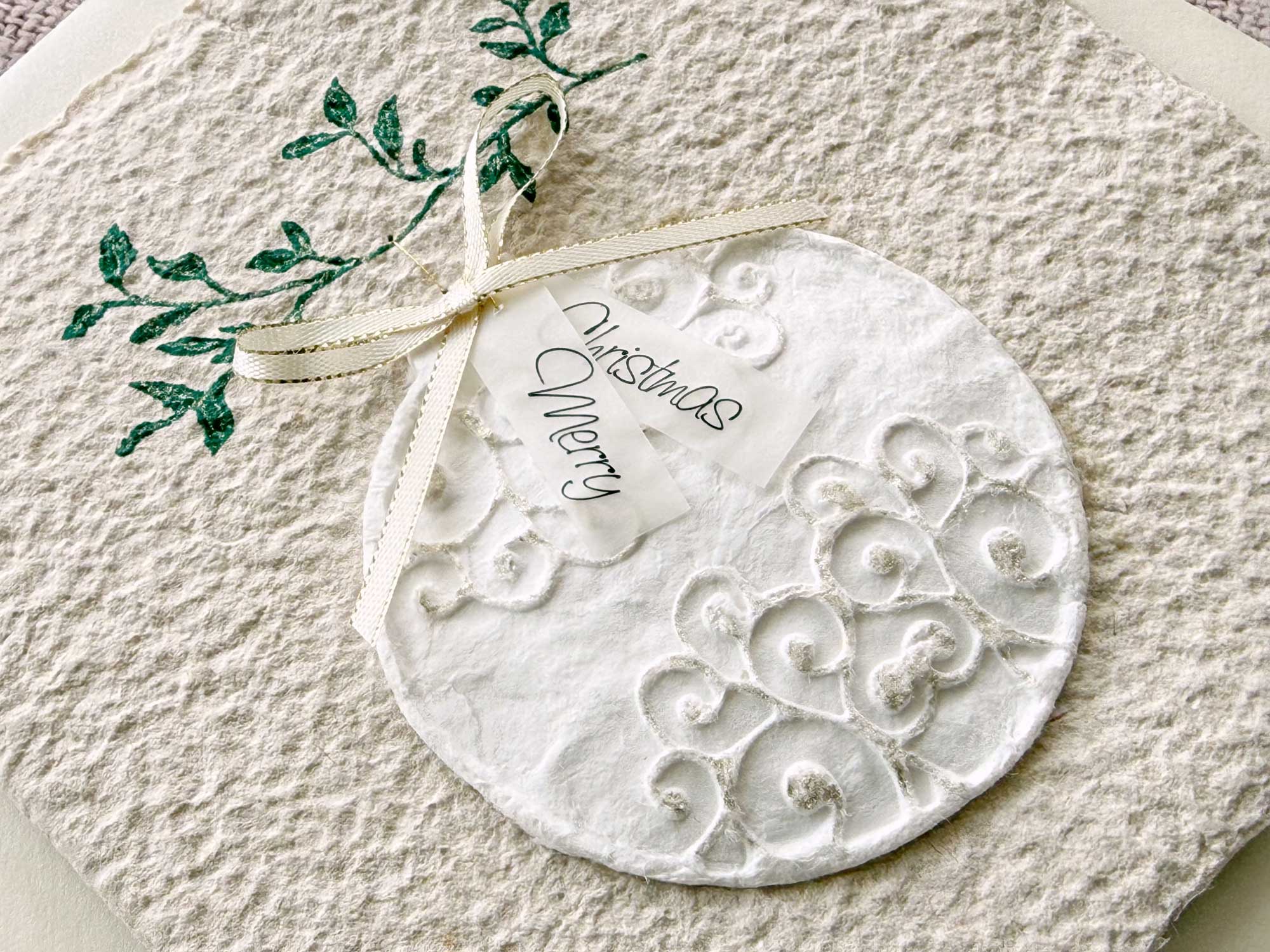 Personalisierbare Weihnachtskarte Christbaumkugel aus handgeschöpftem Papier mit 3D Kugelmotiv, grünem Zweig und cremefarbener Satinschleife.
