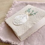 Leicht geöffnete, personalisierbare Weihnachtskarte aus beigem Büttenpapier mit barocker Weihnachtskugel und zart glänzenden Gold Details