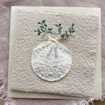 Personalisierbare Weihnachtskarte Weihnachtskugel aus handgeschöpftem Büttenpapier mit reliefartigem Kugelmotiv, grünem Zweig und cremefarbener Satinschleife.