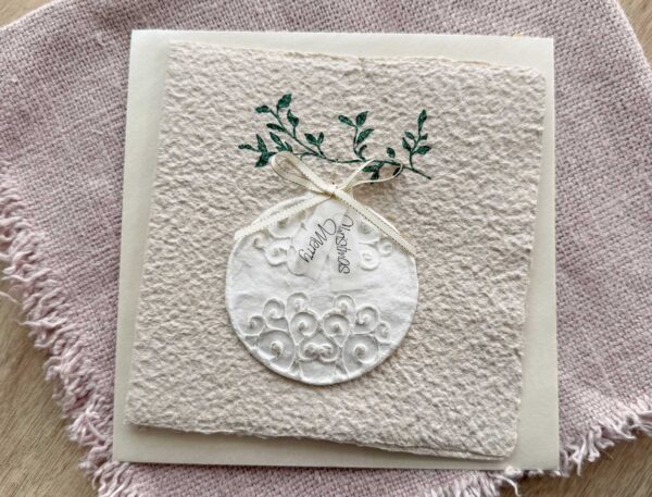 Personalisierbare Weihnachtskarte Weihnachtskugel aus handgeschöpftem Büttenpapier mit reliefartigem Kugelmotiv, grünem Zweig und cremefarbener Satinschleife.